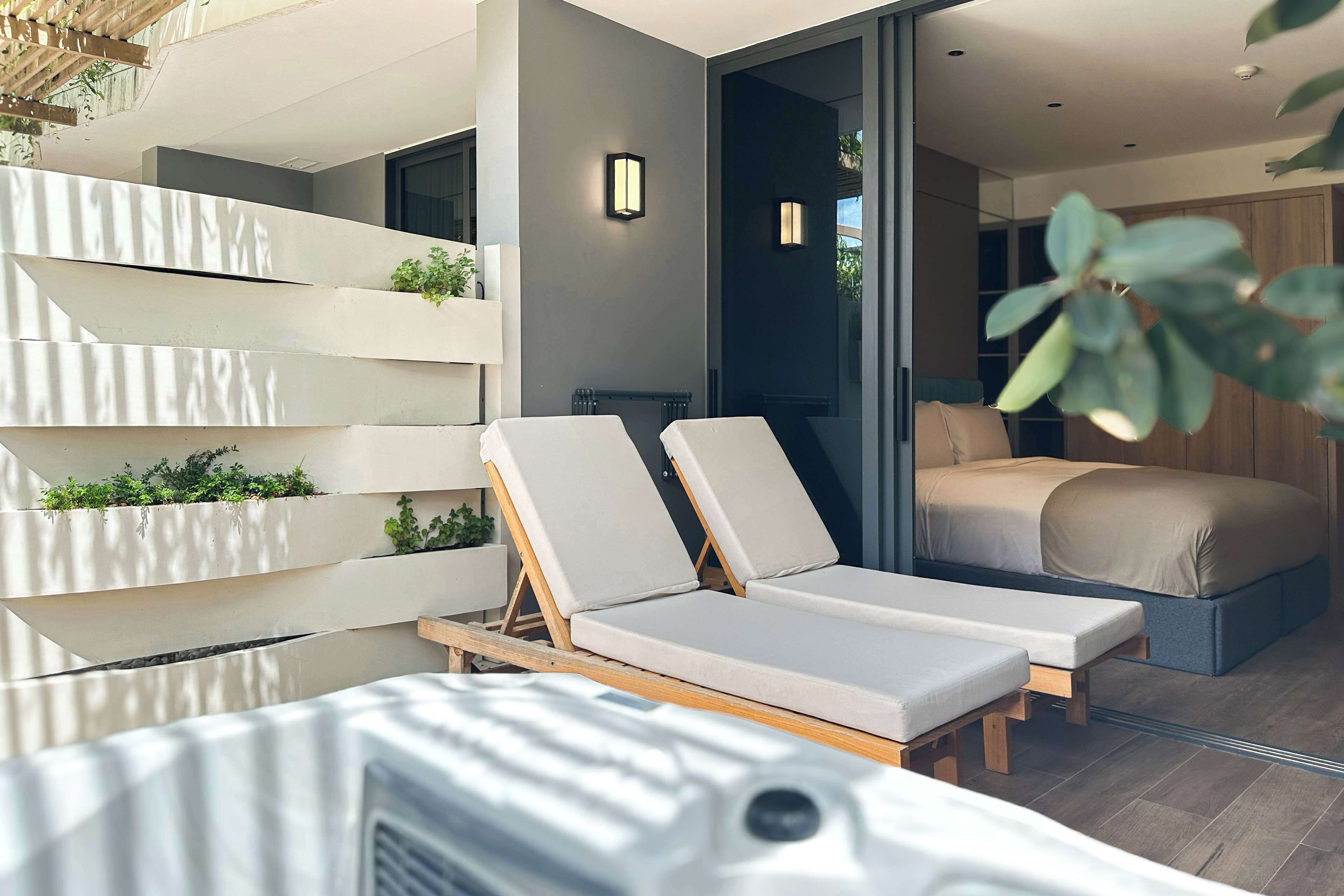Un dormitorio moderno con una cama, un pequeño balcón con plantas y una zona de estar con dos sillones.