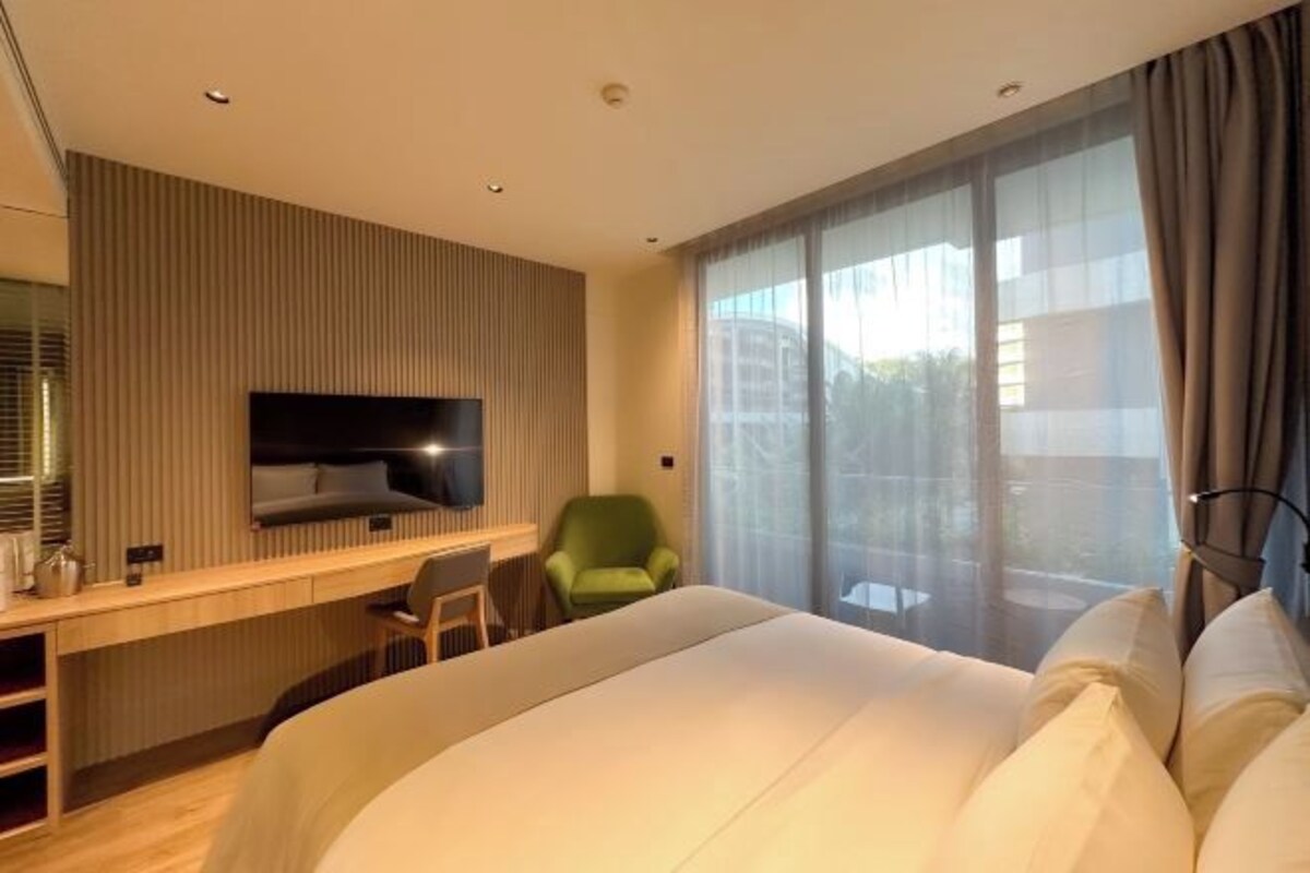 A modern hotel room with a large bed, a desk with a chair, a television, and a large window with curtains.
