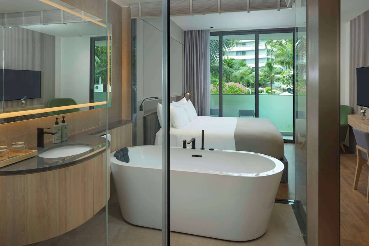 A modern hotel room with a curved bathtub, a large mirror, a flat-screen TV, and a balcony with a view of palm trees.