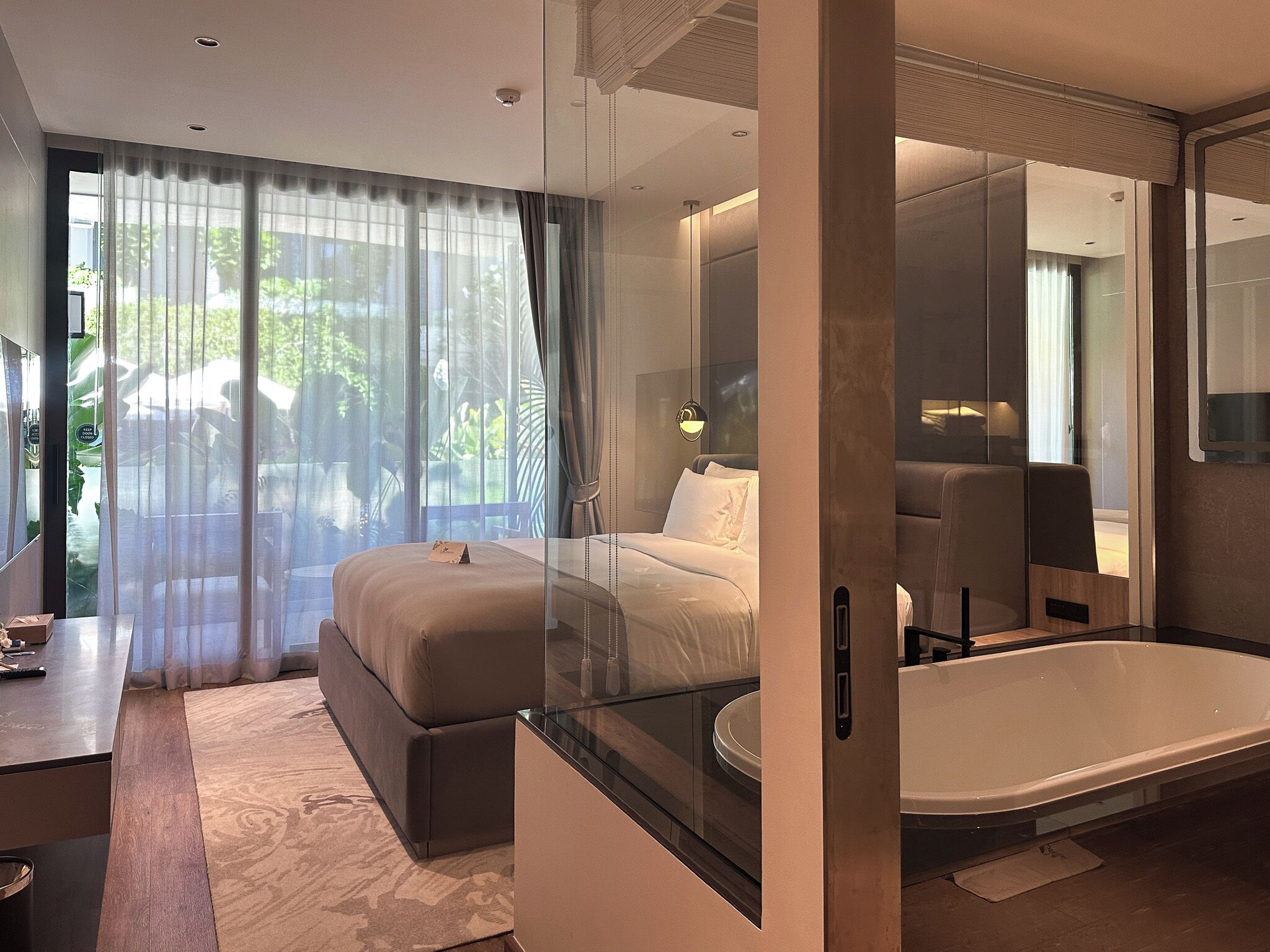 A modern hotel room with a glass-enclosed bed, a large window with sheer curtains, a separate sitting area with a small table, and a standalone bathtub.