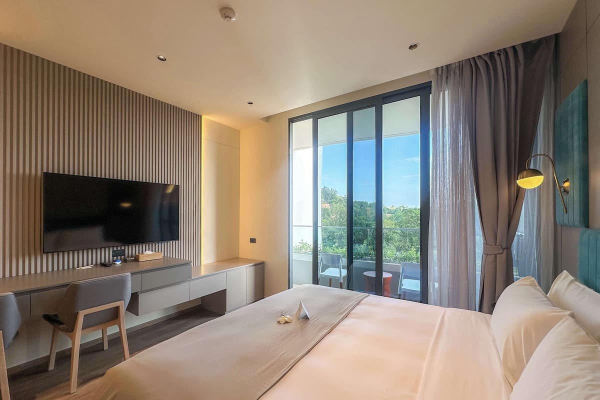 A modern hotel room with a large bed, a desk with a flat-screen TV, and a balcony with a view of greenery.