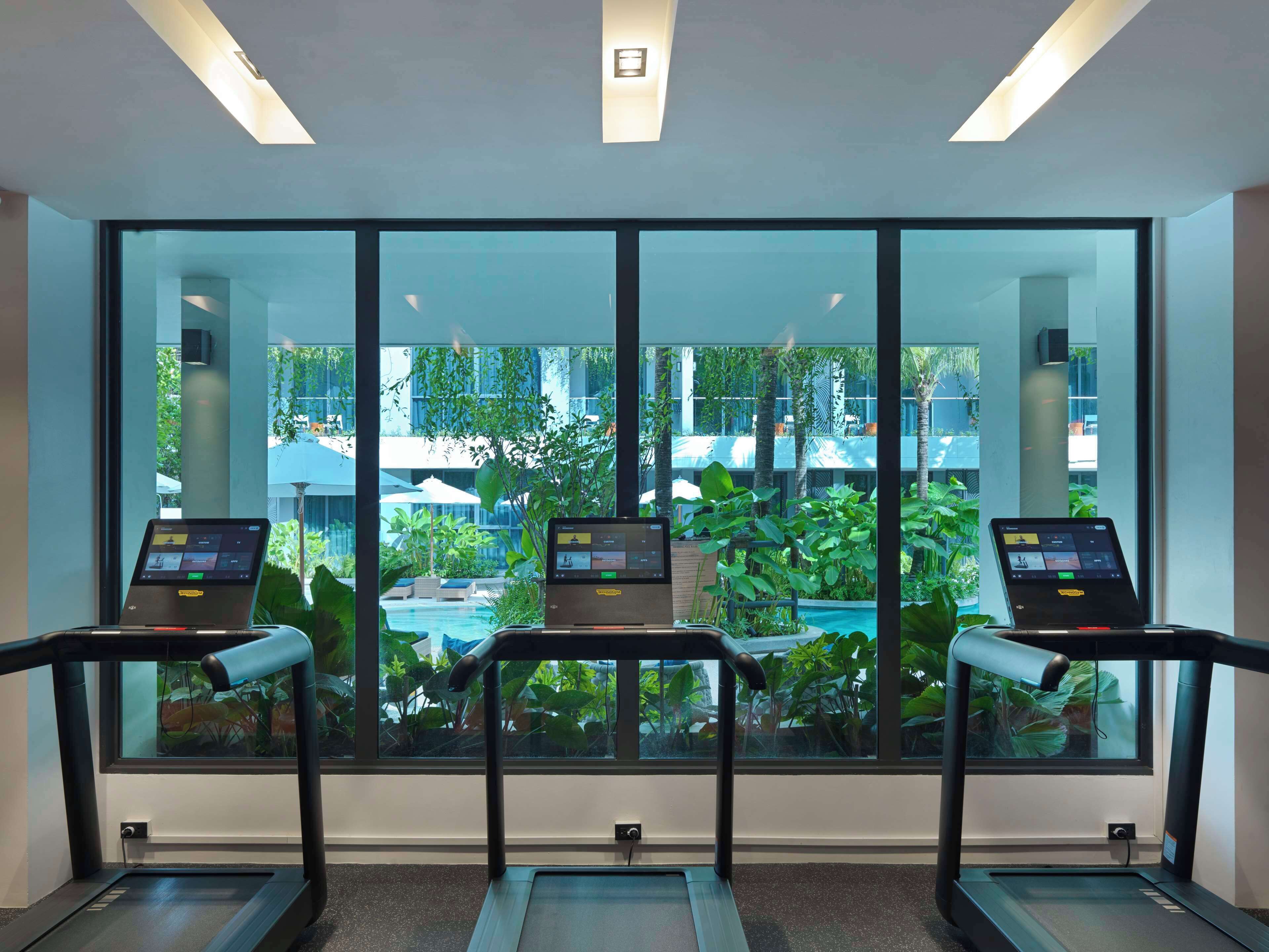 Un área de fitness moderna con cintas de correr, cada una equipada con una computadora portátil que muestra programas de entrenamiento. Amplias ventanas ofrecen vistas a una piscina exterior y abundante vegetación.