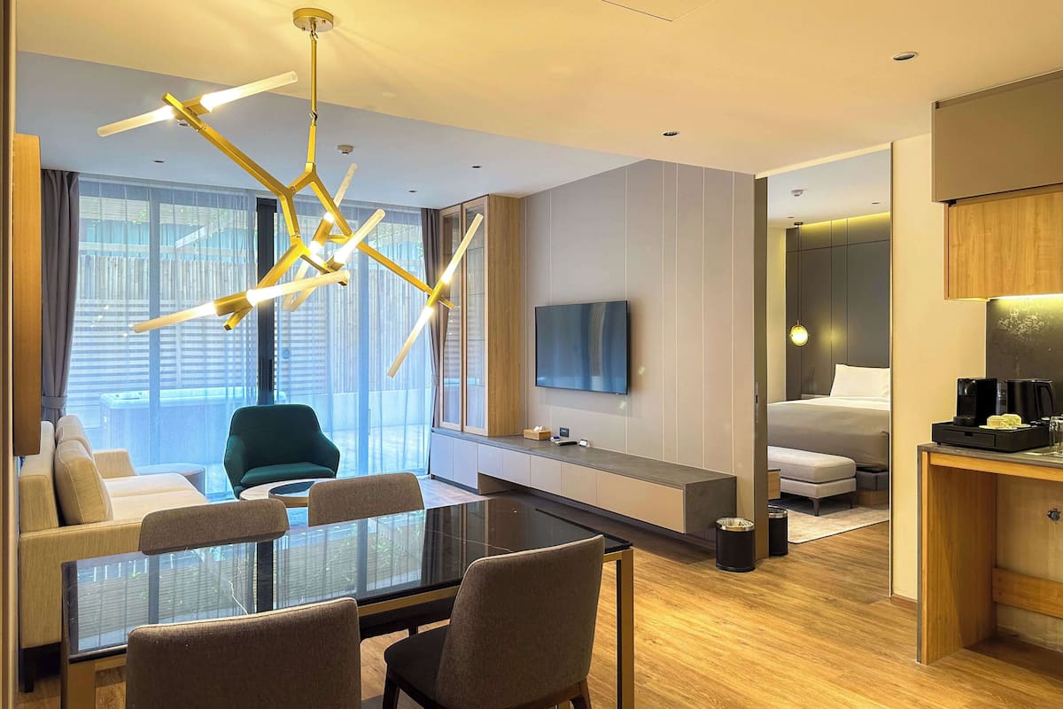 A modern hotel room with a glass dining table, comfortable seating, a TV, a bed, and a coffee machine.