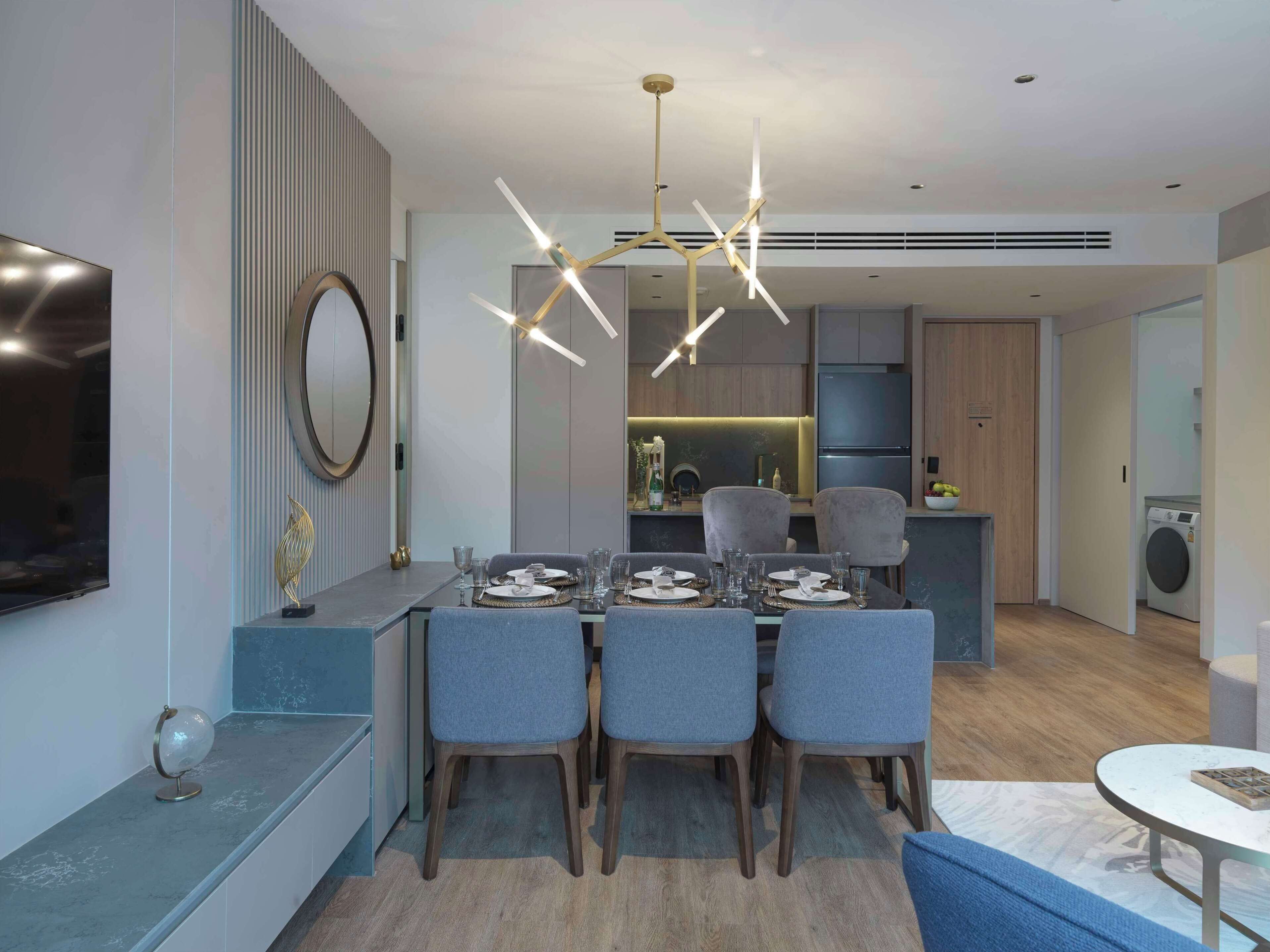 Un moderno comedor con una mesa central dispuesta para cuatro personas, rodeada de sillas tapizadas en gris. Un candelabro contemporáneo cuelga encima, y un televisor de pantalla plana está montado en la pared a la izquierda.