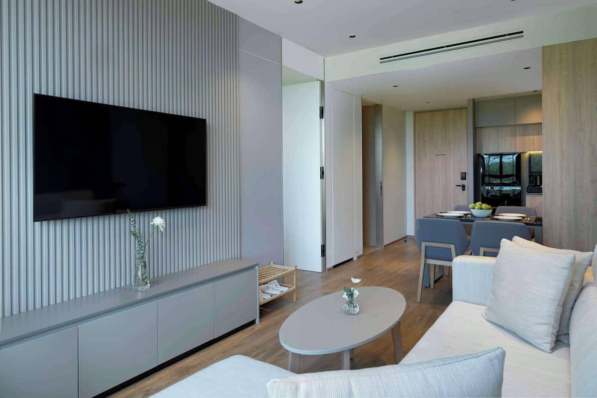 A modern living room with a flat-screen TV mounted on the wall, a sofa, a coffee table, and a kitchen area visible through an open door.