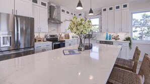 Une cuisine moderne avec des meubles blancs, un grand îlot central, des appareils en acier inoxydable, des luminaires suspendus et des chaises en osier.