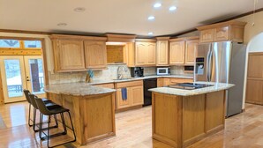 A spacious kitchen with wooden cabinets, a stainless steel refrigerator, a microwave, and a dishwasher. There is a bar stool and a window with a view of the outside. - Relaxing Lake Hamilton Home with Private Dock, Hot Tub, Kayaks, Fire Pit (Royal)