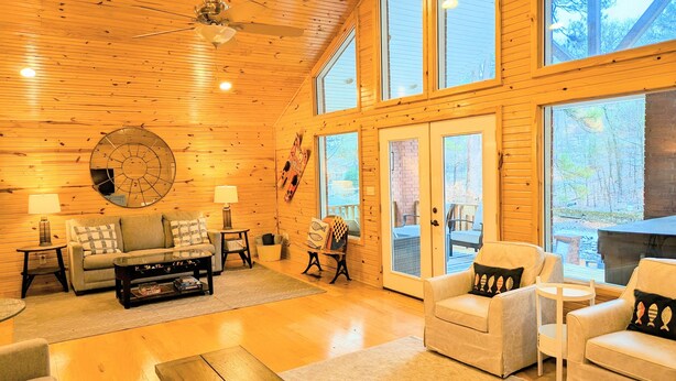 A spacious living room with wooden floors, a ceiling fan, a large round mirror, and a view of the outdoors through large windows. - Relaxing Lake Hamilton Home with Private Dock, Hot Tub, Kayaks, Fire Pit (Royal)