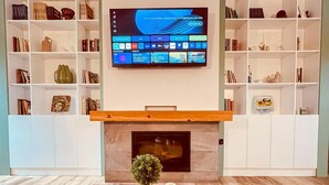 Un salon avec un téléviseur fixé au mur, une cheminée et des étagères remplies de livres et d’objets décoratifs.
