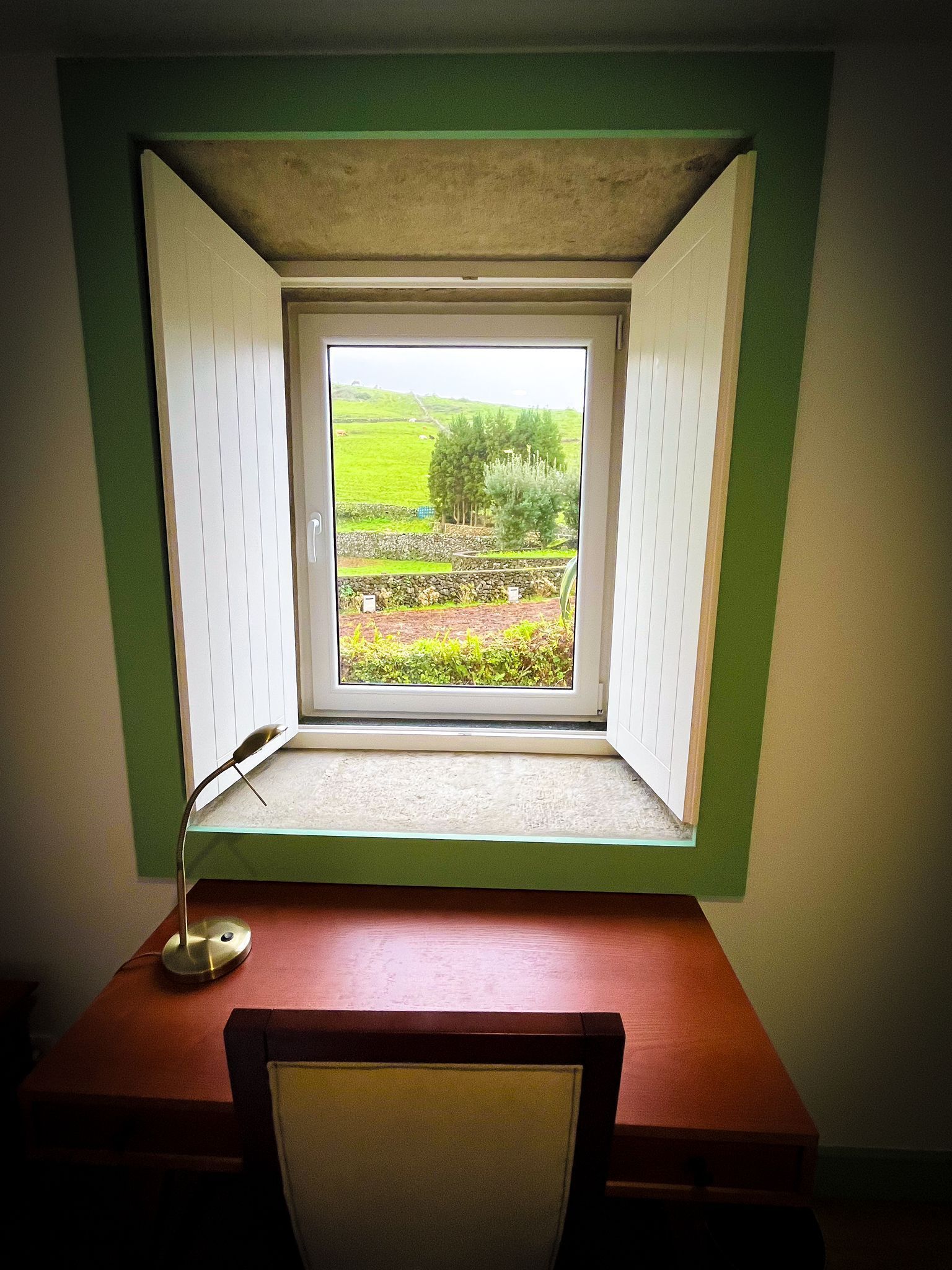 Una habitación con un escritorio, una silla y una ventana con un contraventanas abierta.