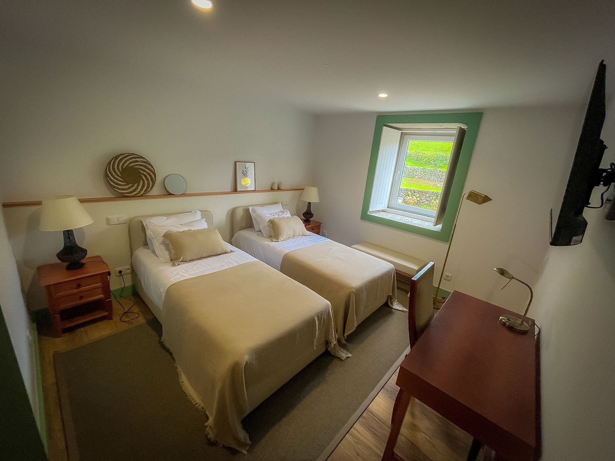 A bedroom with two beds, a small table, a window with a view, and a bedside lamp.