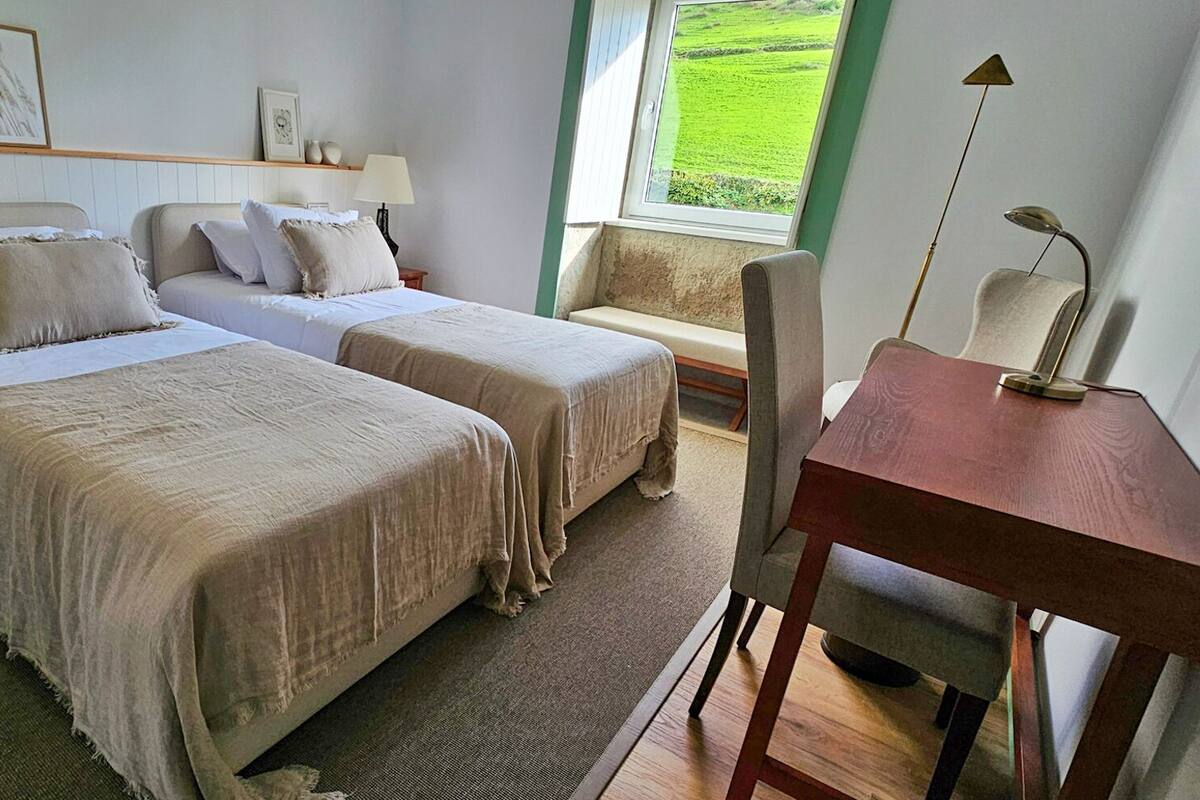 A bedroom with two beds, a desk, and a chair. There is a window with a view of greenery outside.