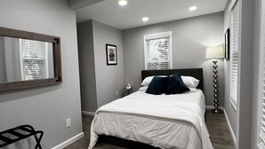 A bedroom with a bed, a bedside table, a lamp, a window with blinds, and a mirror. - Peaceful Riverfront Cottage with Private Hot tub (Lake Katrine)