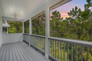A covered porch with a view of a forested area, a ceiling fan, and a small structure with a railing.