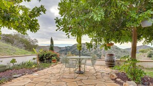 Un patio de piedra con una mesa redonda y sillas, rodeado de follaje y vistas a las colinas.