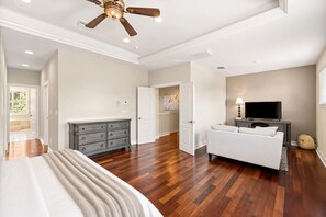 A spacious bedroom with hardwood flooring, a large bed, a dresser, a ceiling fan, a television, and a door leading to another room.
