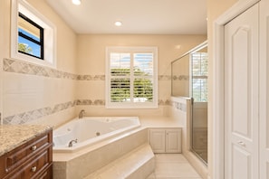 A bathroom with a large bathtub, a shower area, and a window with blinds.