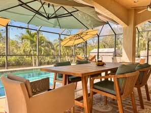 A poolside area with a wooden table and chairs, a yellow umbrella, and a view of the surrounding greenery.
