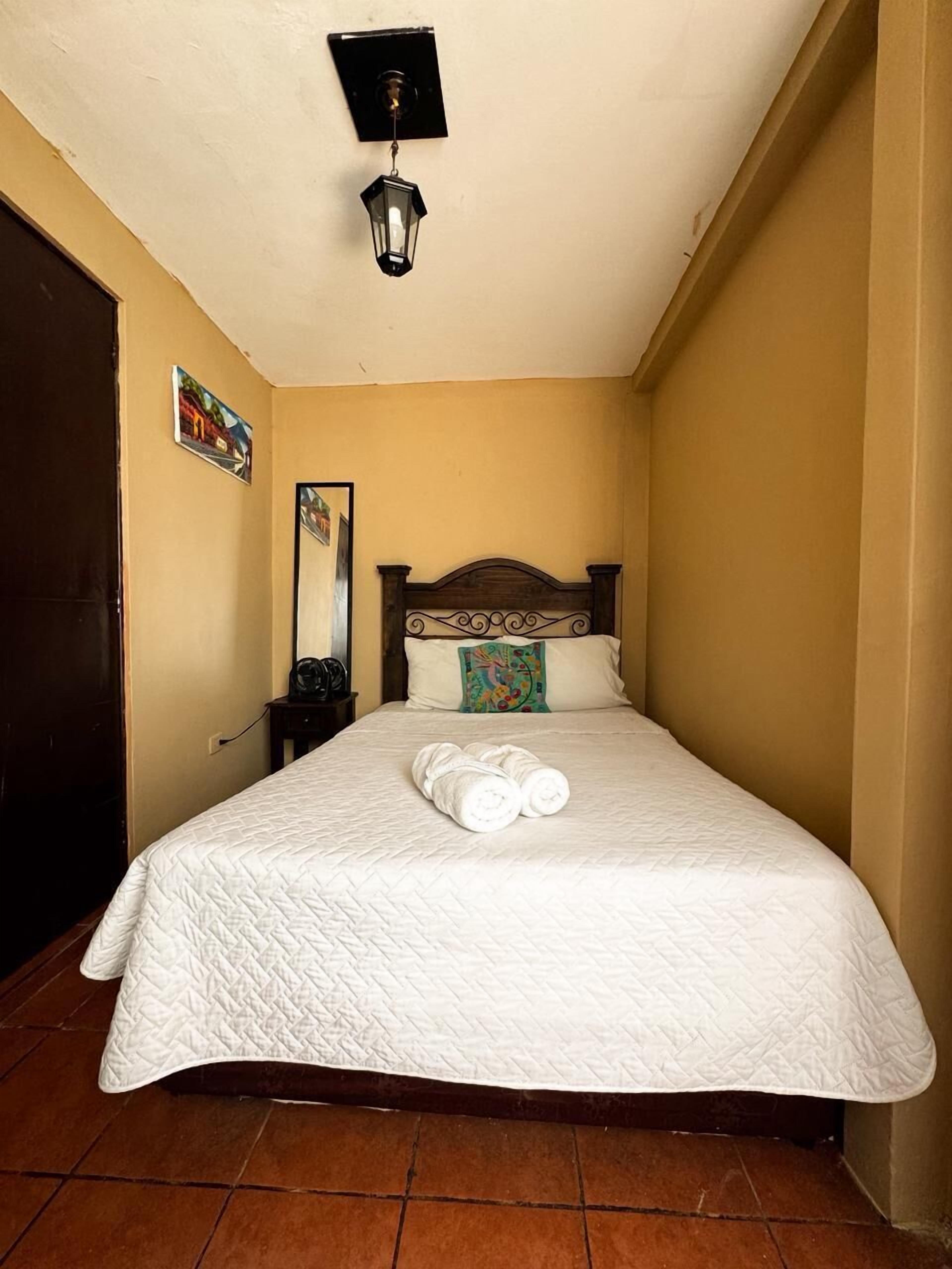 A neatly made bed with a white quilted bedspread and rolled towels, a wooden headboard, a wall-mounted light fixture, and a door to the left.
