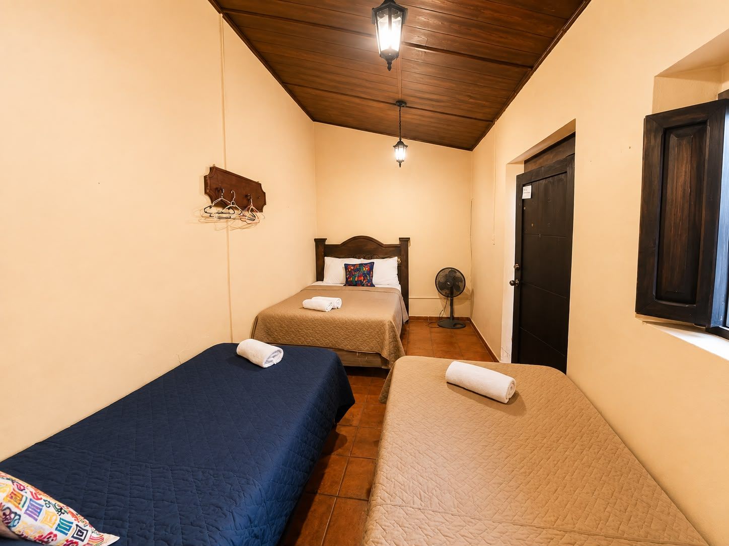 Ein Hotelzimmer mit zwei Einzelbetten, einer Holzbalkendecke, einem Wandventilator und einer Tür mit Fenster.