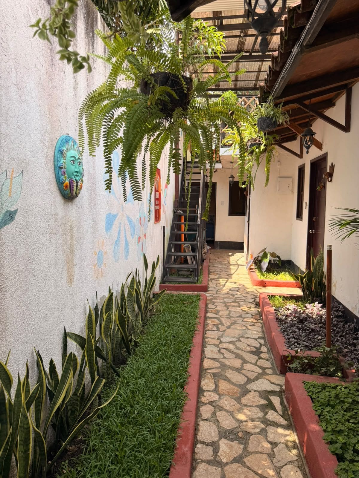 Un estrecho sendero de piedra bordeado por follaje y un mural, que conduce a un edificio con una escalera de madera.
