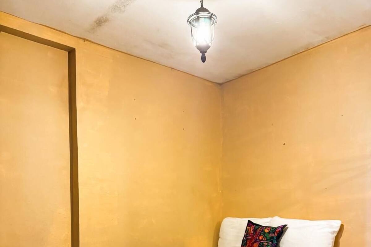 A single bed with a blue bedspread, a white pillow, and a colorful pillow on a wall with a light fixture above.