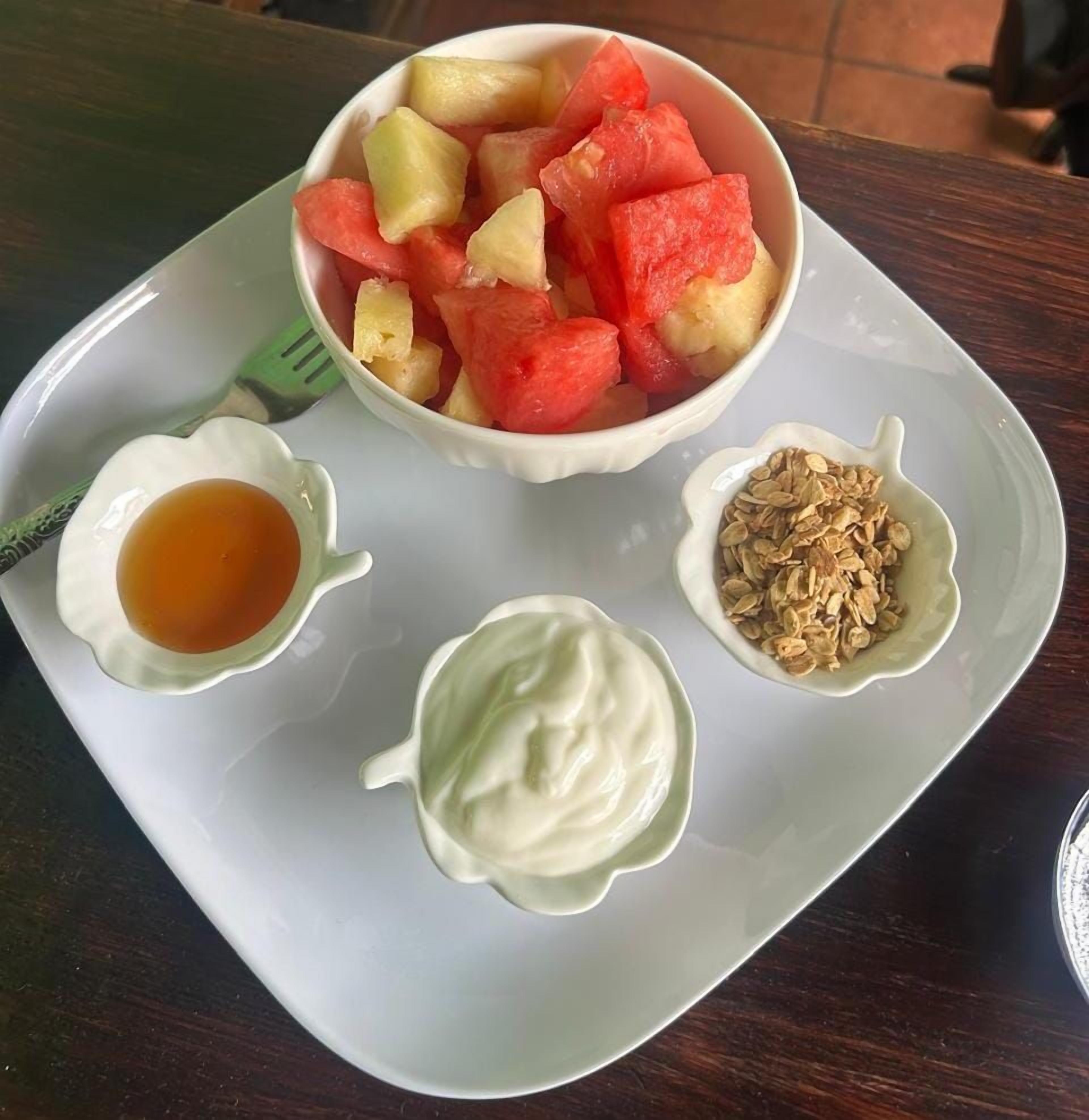 Een bord met een schaal fruit, een klein schaaltje honing, een klein schaaltje yoghurt en een klein schaaltje granola.