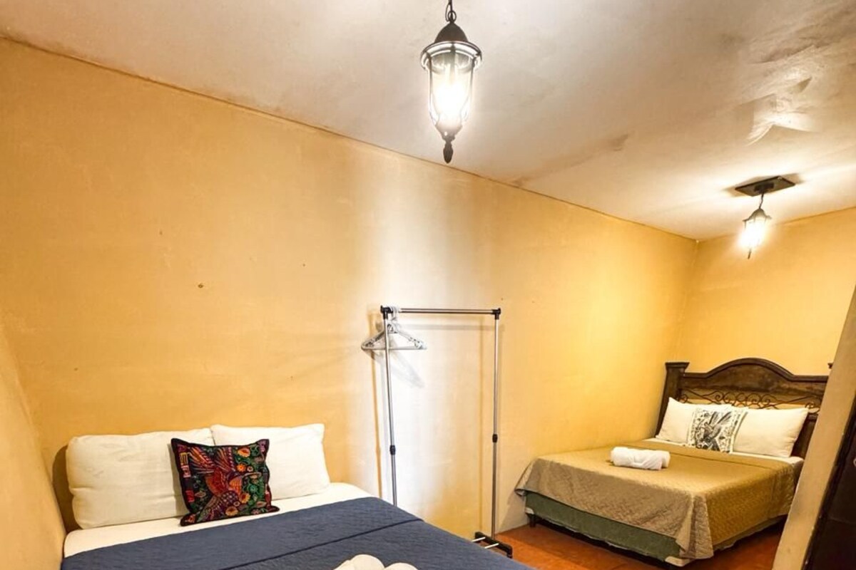 Un lit simple avec une couvre-lit bleue et deux serviettes enroulées, une table de chevet, un luminaire suspendu et une petite table avec une chaise dans un coin.