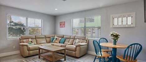 A living room with a beige sectional sofa, a wooden coffee table, and a dining area with blue chairs and a wooden table.