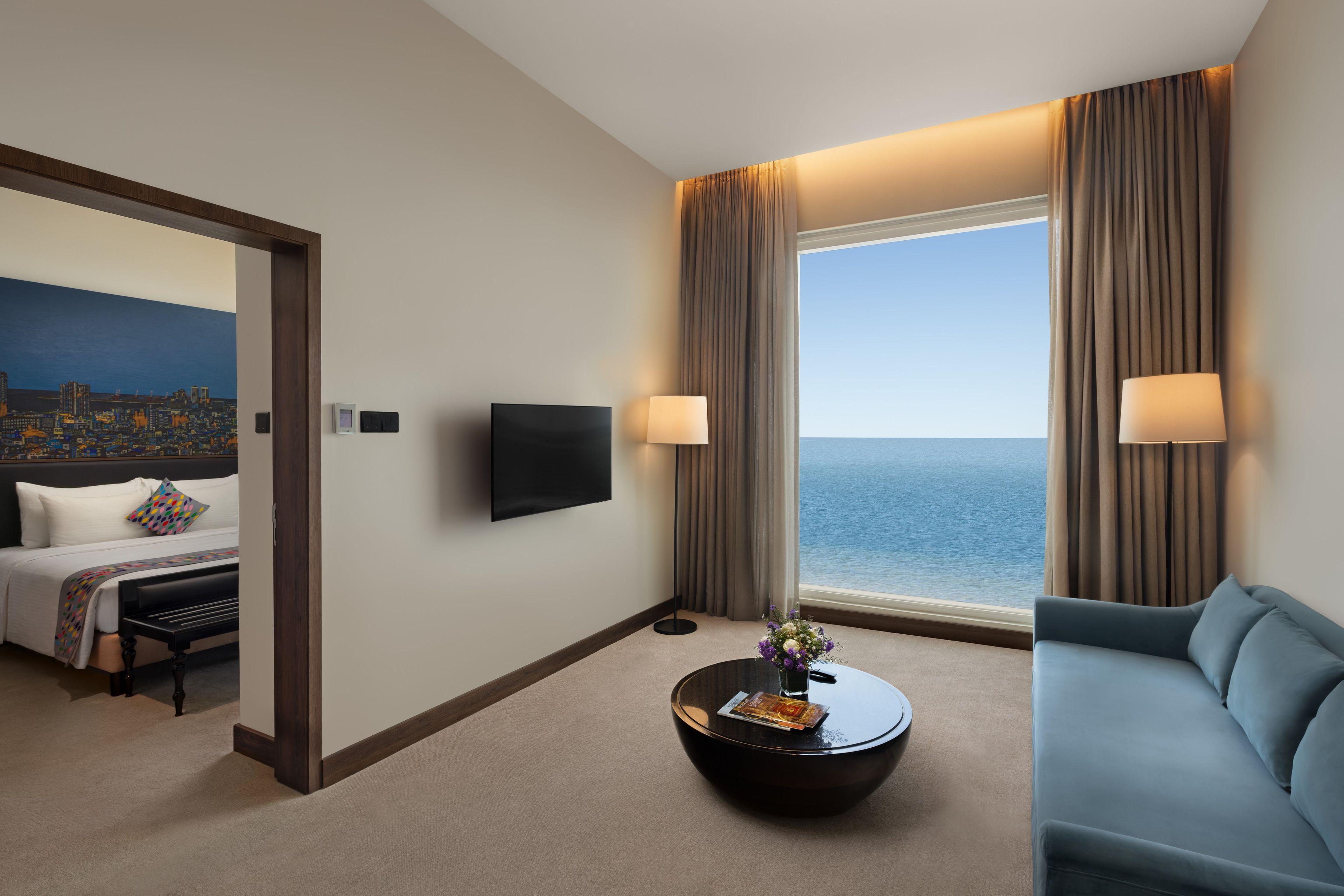 A hotel room with a bed, a sofa, a coffee table, a TV, and a view of the sea.