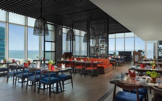 Un restaurante moderno con techos altos, ventanas amplias y vista al mar. Las mesas están preparadas con servilletas rojas y plantas en macetas.