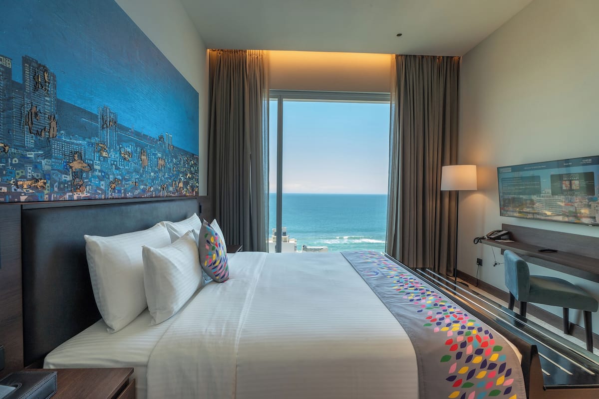 A hotel room with a large bed, a desk, a chair, a TV, and a view of the ocean.