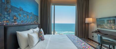A hotel room with a large bed, a desk, a chair, a TV, and a view of the ocean.