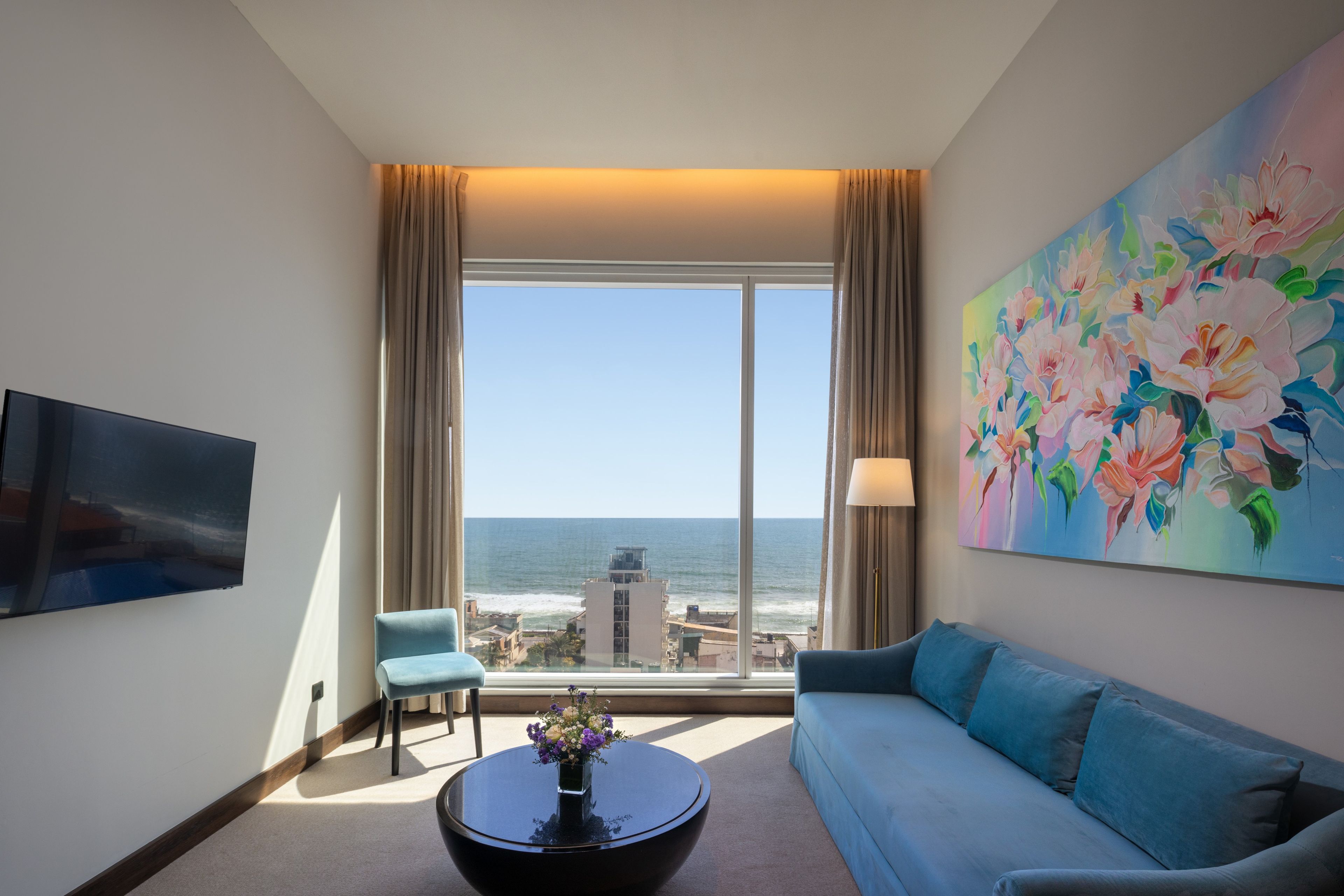 Habitación de hotel con un ventanal grande, un sofá, una silla, una mesa de centro y un cuadro.