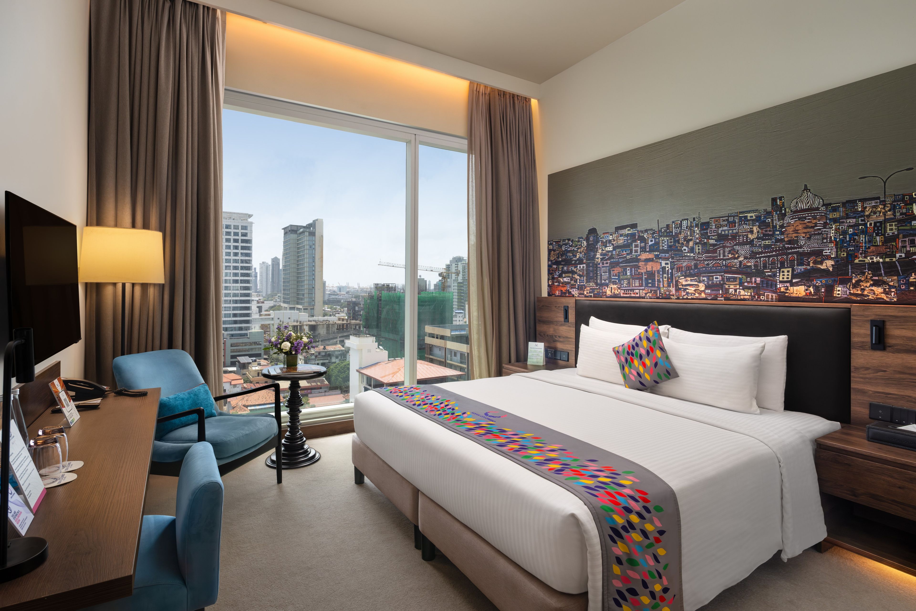 Habitación de hotel con una cama grande, un escritorio, una silla, un televisor y vistas a la ciudad.