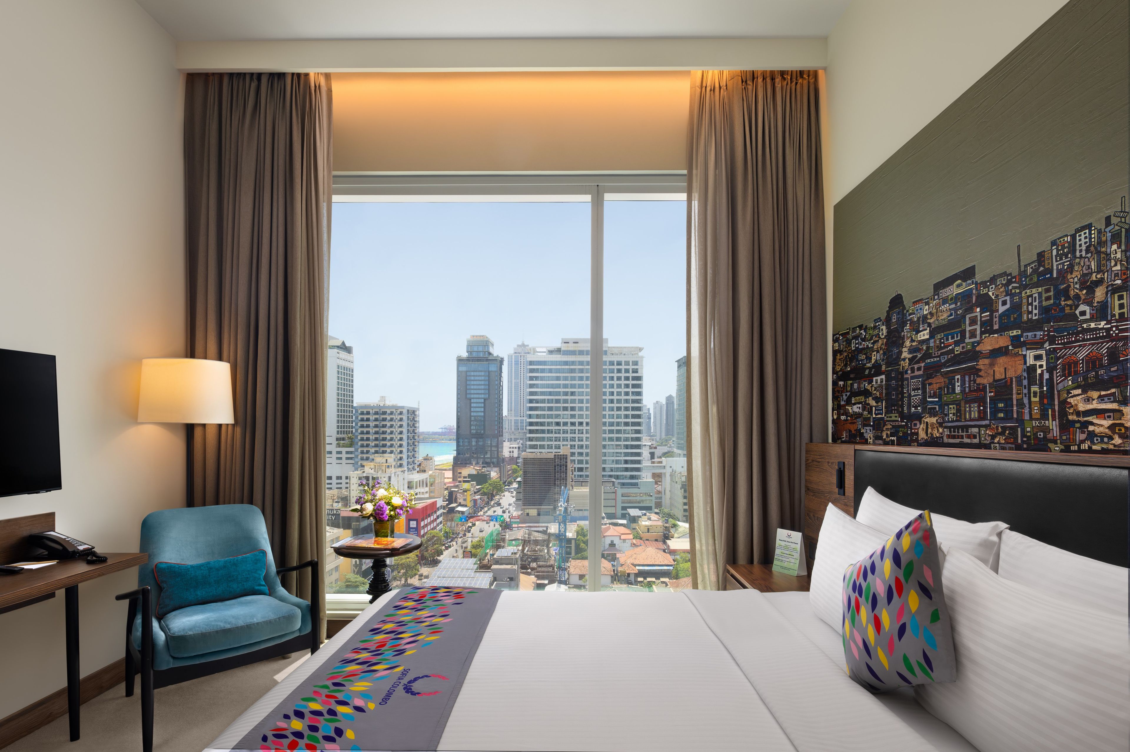 A hotel room with a large bed, a chair, a desk, a lamp, and a view of a cityscape through large windows.