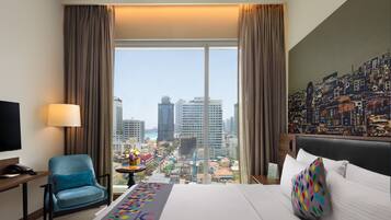 A hotel room with a large bed, a chair, a desk, a lamp, and a view of a cityscape through large windows.