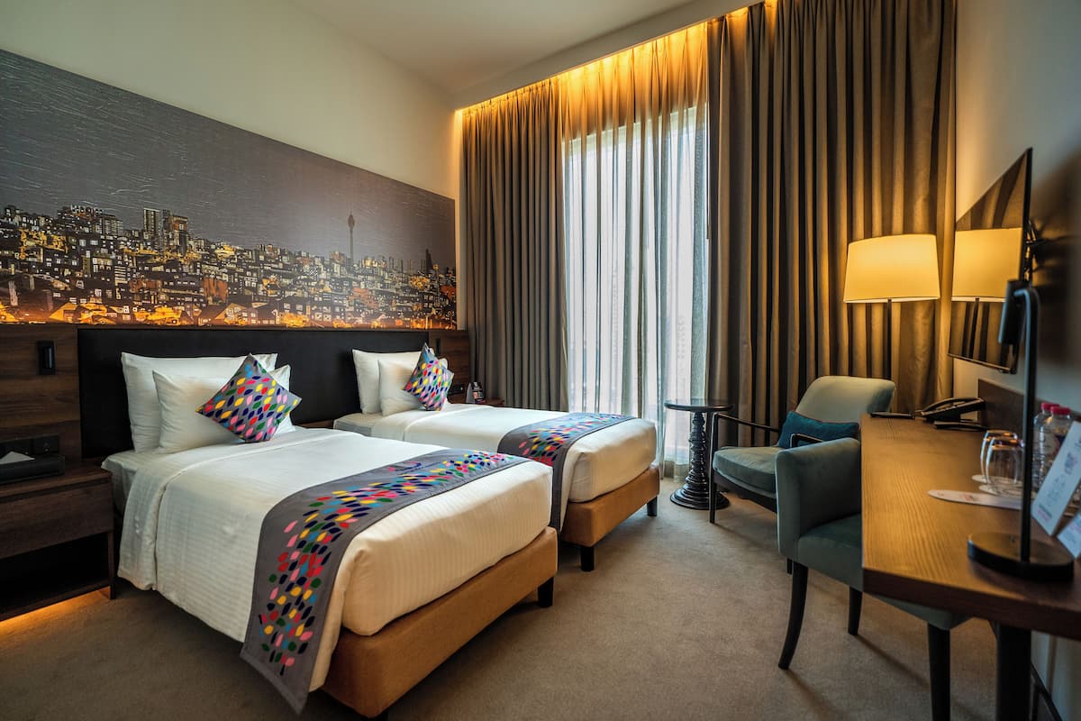 A hotel room with two beds, a desk, a chair, a cityscape painting, and a large window with curtains.