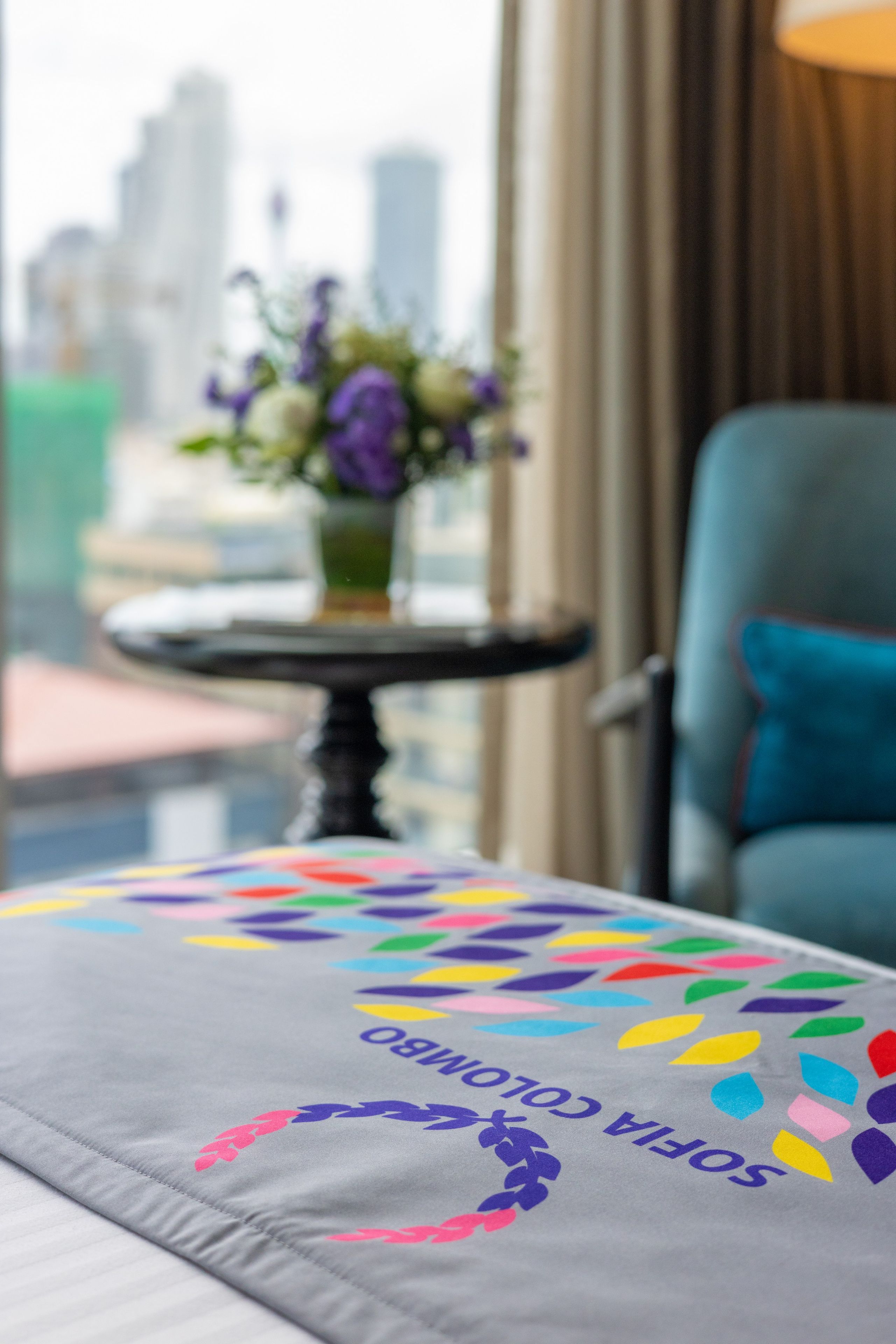 A hotel room with a view of the cityscape, a table with a colorful tablecloth, and a chair with a blue cushion.
