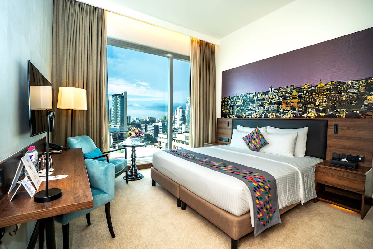 A hotel room with a large bed, a desk, a chair, and a cityscape view through the window.