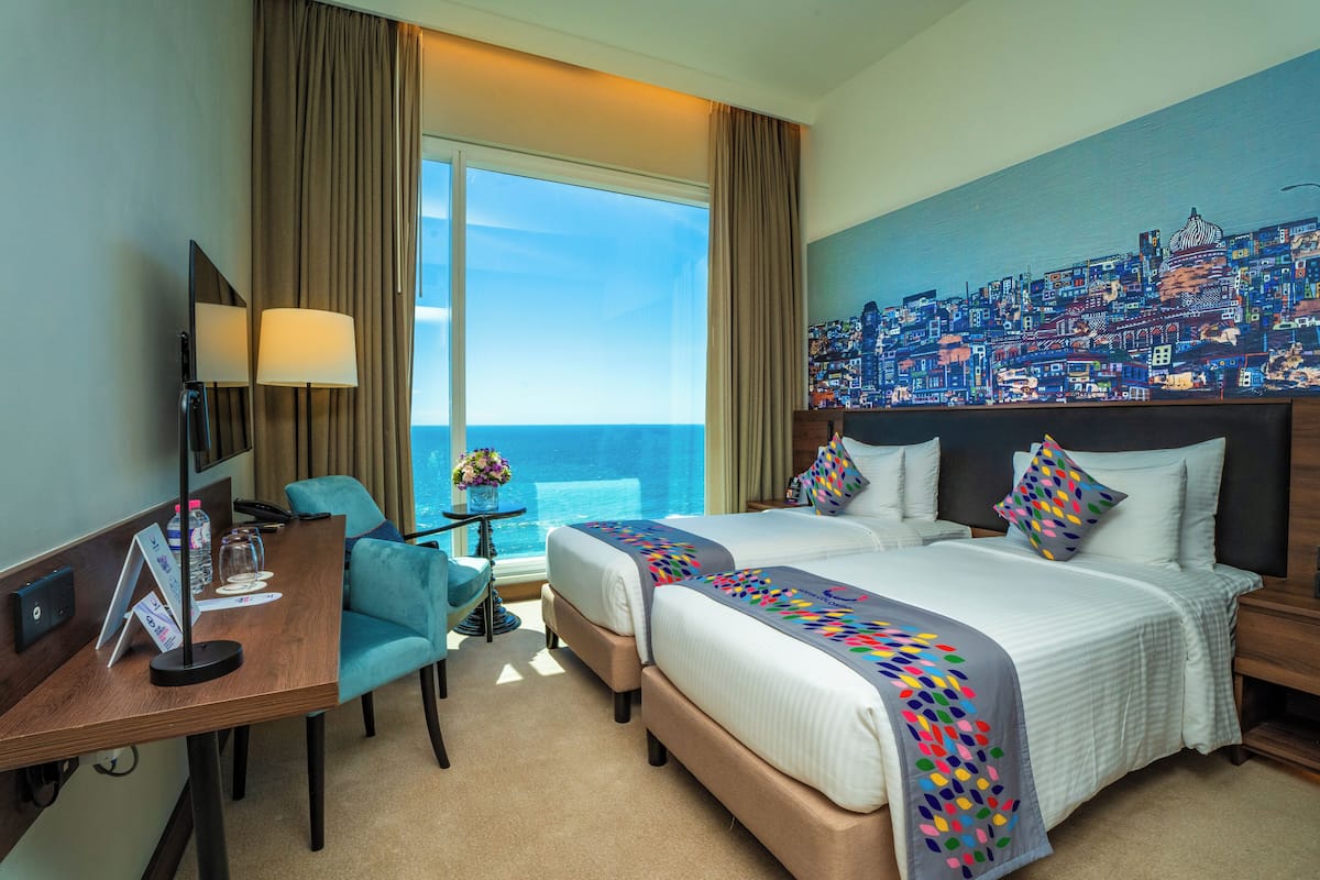 A hotel room with two beds, a desk, a chair, a TV, and a large window with a cityscape view.