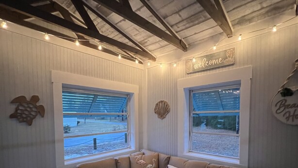 A cozy room with a sectional sofa, two windows with blinds, a ceiling with exposed beams, and a welcome sign.