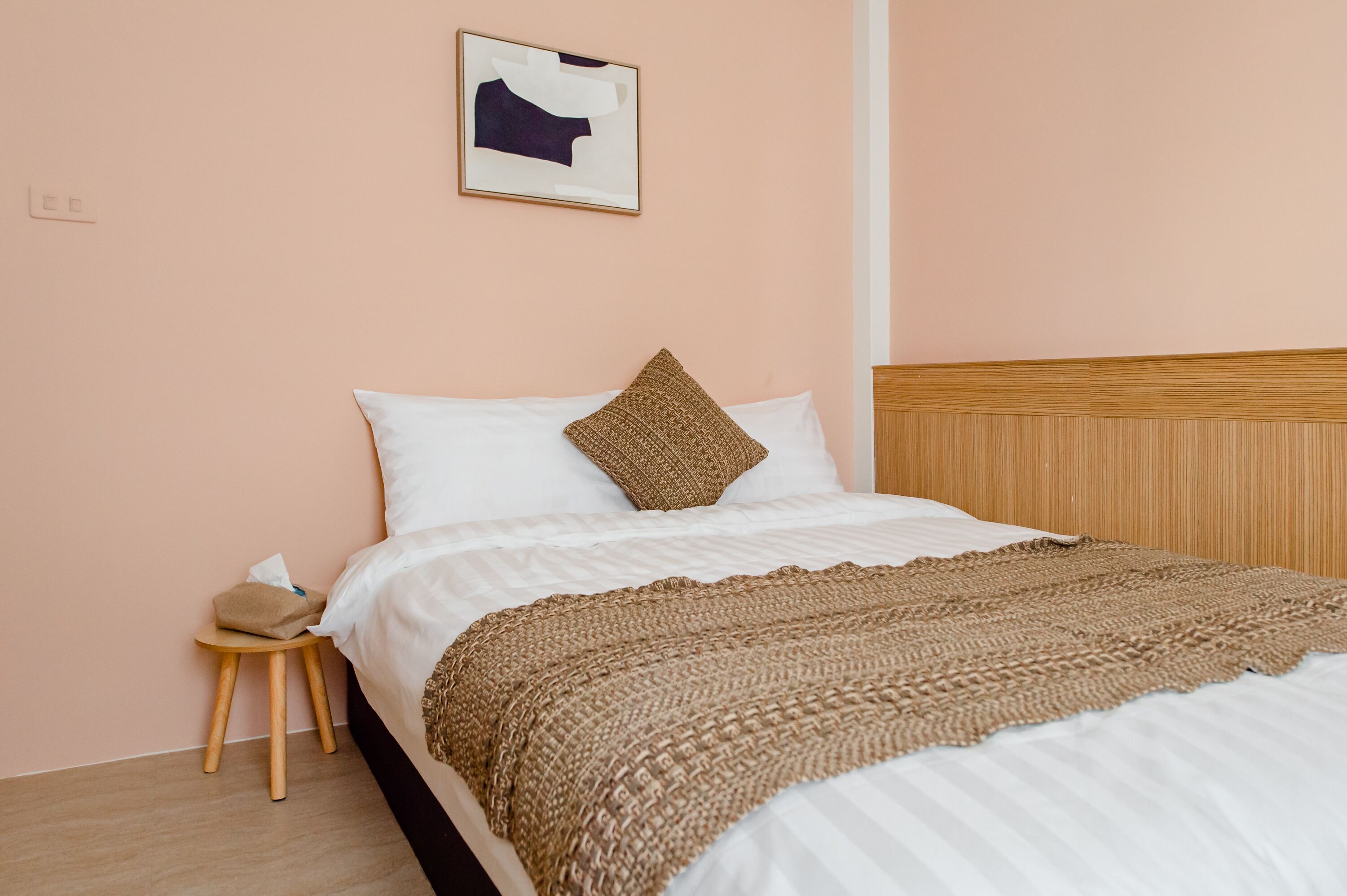 A neatly made bed with a brown knitted blanket and a white pillow, a small wooden stool with a tissue box beside it, and a framed abstract artwork on the wall.
