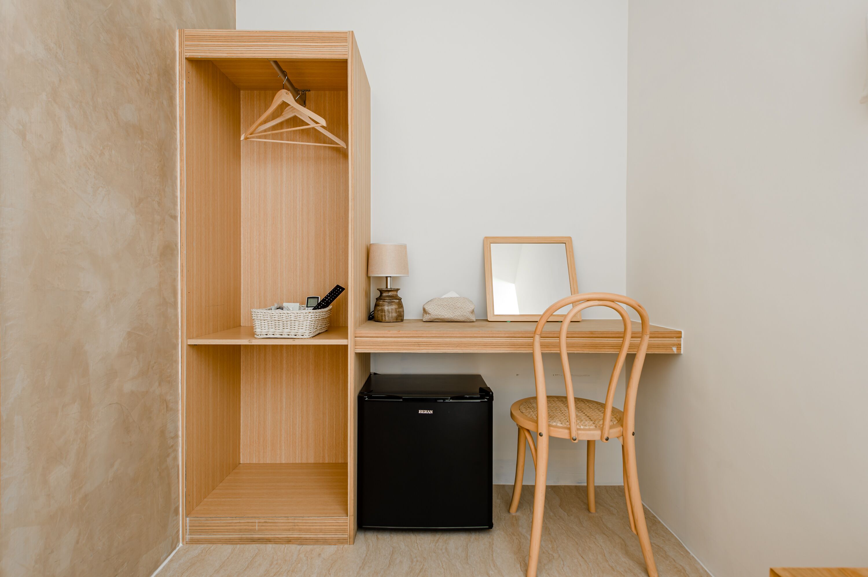 A compact room with a wooden wardrobe, a desk with a chair, a small black refrigerator, and a hanging light fixture.