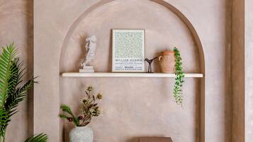 Una obra de arte en un marco sobre una estantería, un jarrón blanco con flores secas, una planta pequeña en maceta y un dispositivo electrónico negro.