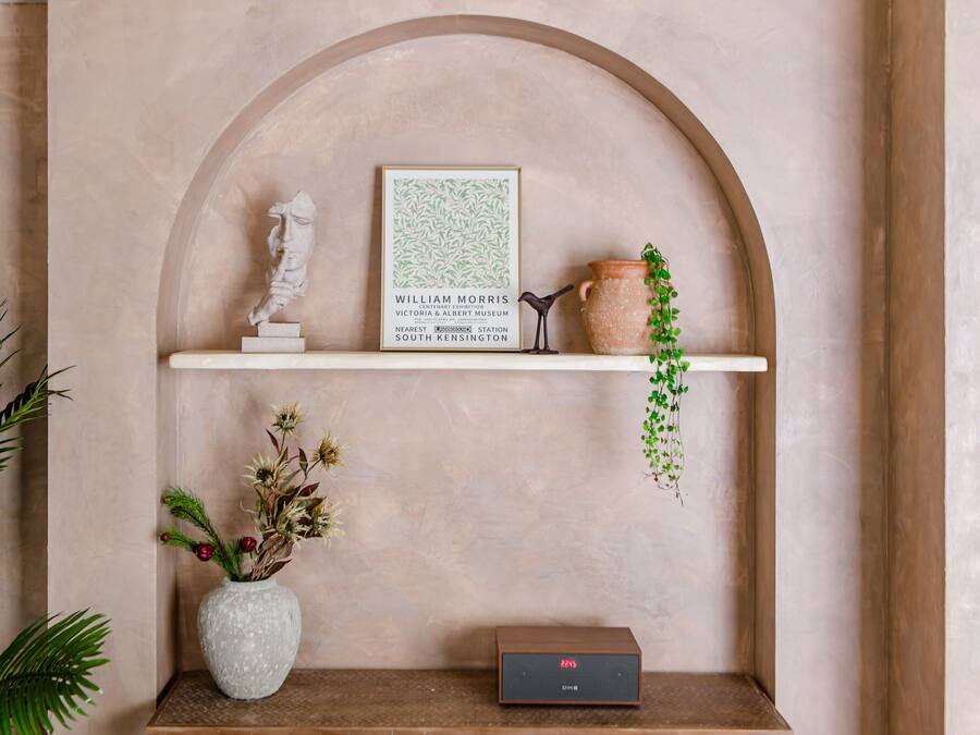 Una obra de arte en un marco sobre una estantería, un jarrón blanco con flores secas, una planta pequeña en maceta y un dispositivo electrónico negro.