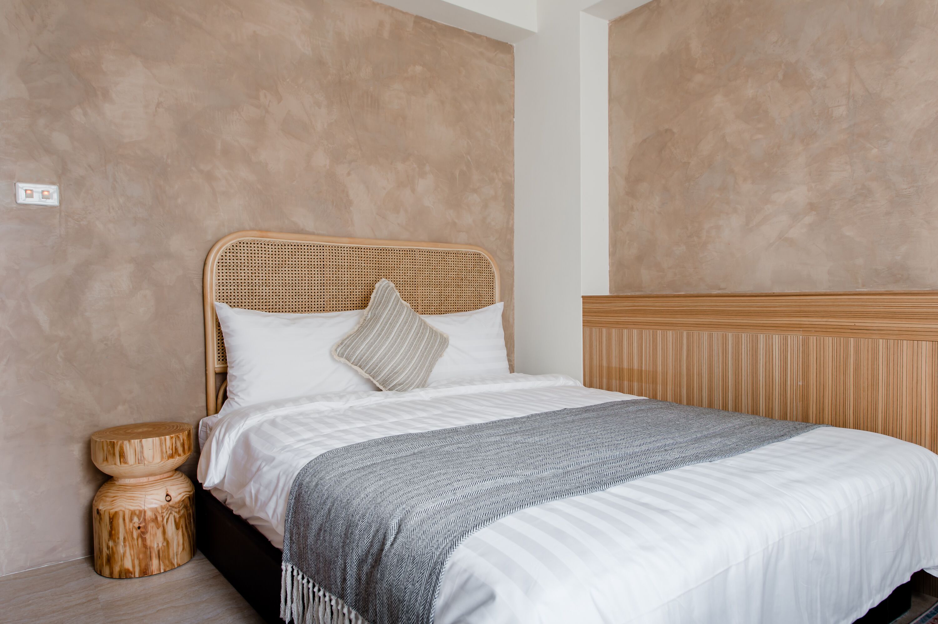 Un letto rifatto con una coperta a righe, una testiera intrecciata e un comodino in legno con uno sgabello rotondo di legno accanto.