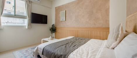 A bedroom with a bed, a bedside table with a plant, a wall-mounted TV, and an air conditioning unit.