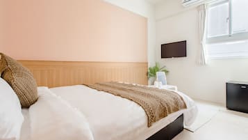 A hotel room with a bed, a flat-screen TV mounted on the wall, a window with curtains, and a small black refrigerator.