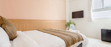 Habitación de hotel con cama, televisor de pantalla plana montado en la pared, ventana con cortinas y un refrigerador negro pequeño.